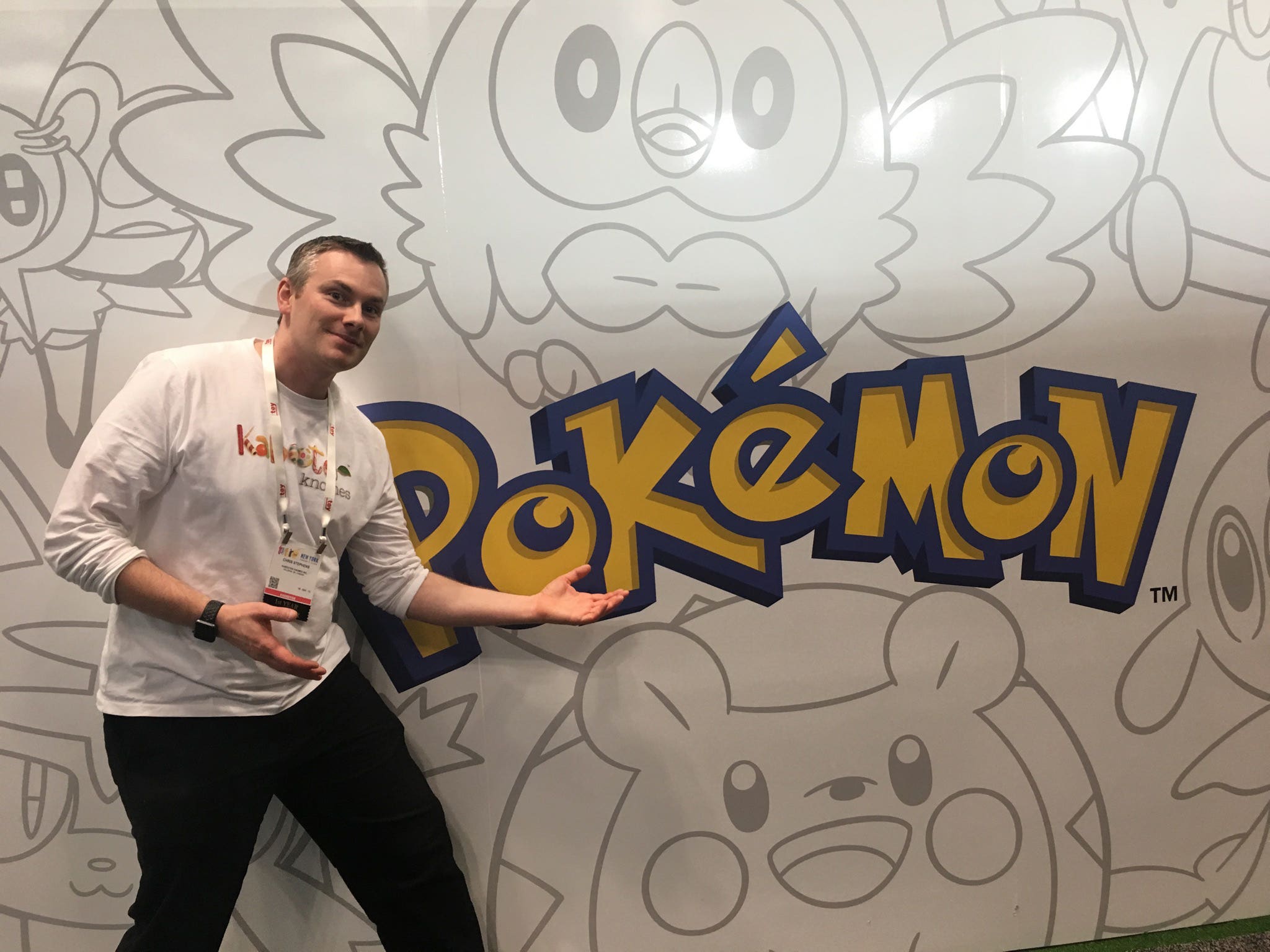 Chris Stephens poses next to a large Pokémon logo at the New York Toy Show, symbolizing the connection between Twirling Umbrellas and popular culture.