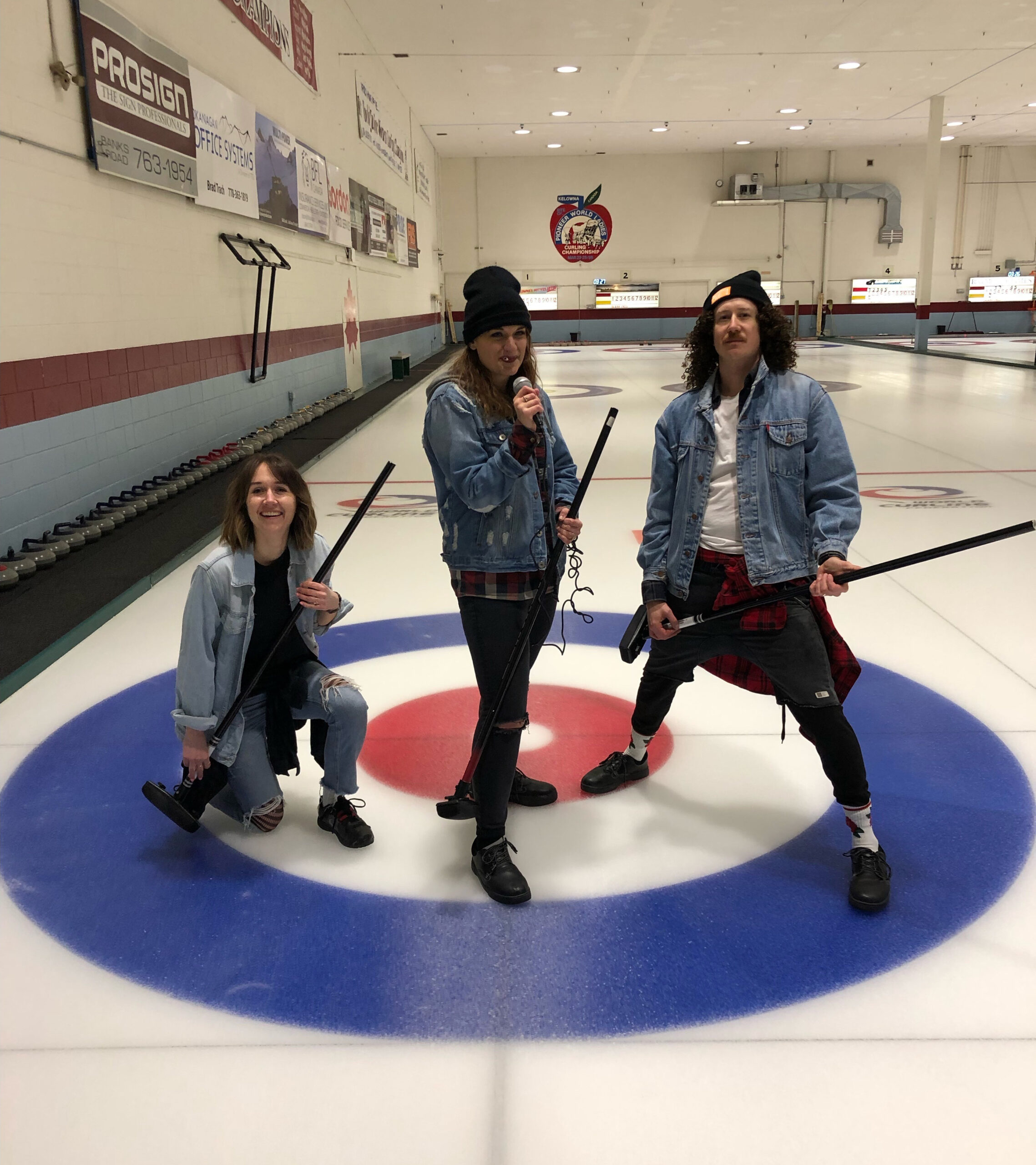 Three Twirling Umbrellas team members on an ice rink with curling brooms, playfully mimicking a rock band pose.