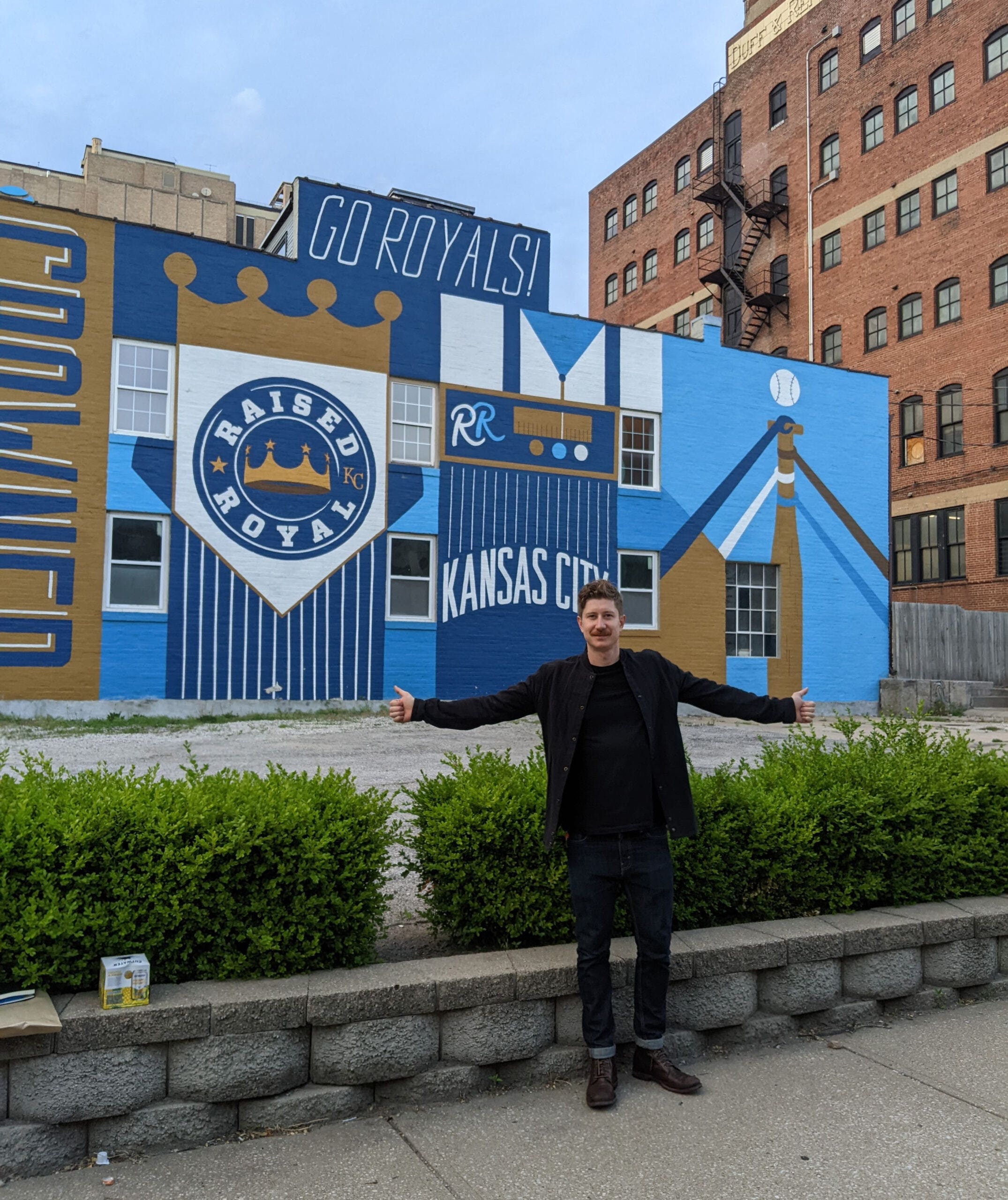 Ryan McGrew stands in front of a Kansas City Royals mural with arms wide open, displaying excitement and team spirit.